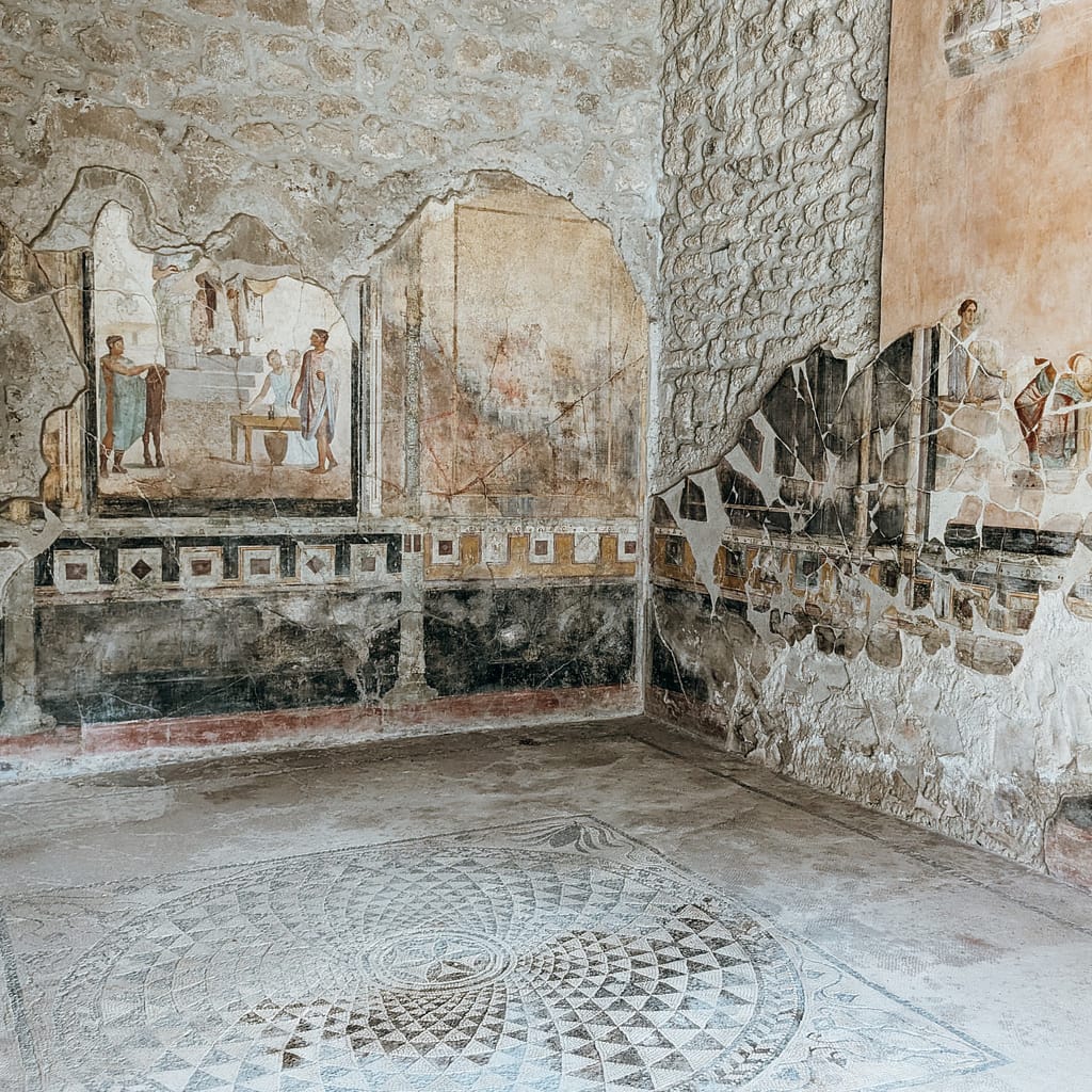 Circle Photo_Pompeii Ancient frescoes and mosaic floor in a partially ruined room with stone walls, depicting people and geometric patterns.