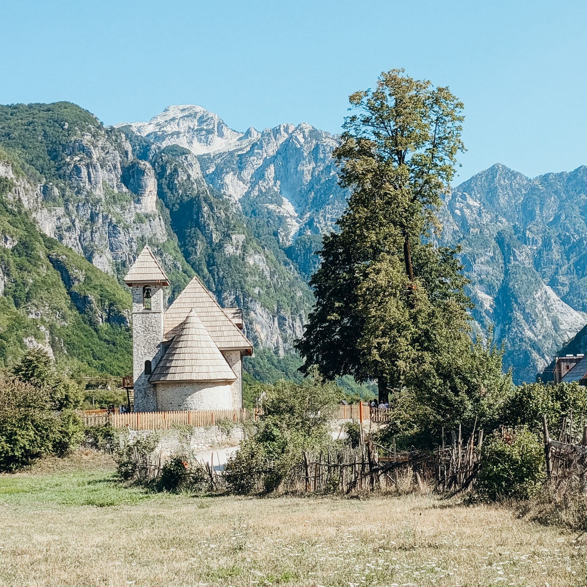 Cirlce A small stone church with a tower, surrounded by trees and mountains, stands under a clear blue sky in a rural landscape.